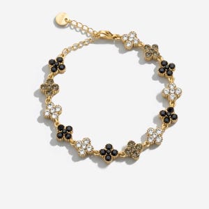 Op de afbeelding: Een goudkleurige kettingarmband met een reeks afwisselende klaverblad-vormige bedels. De bedels zijn ingelegd met heldere, zwarte en rookgrijze kristallen. De armband is voorzien van een karabijnsluiting en een verstelbare verlengketting.