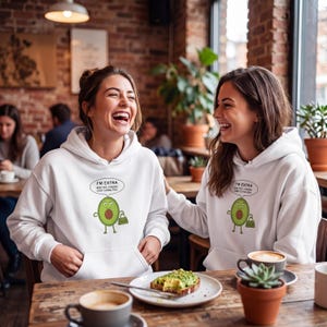 Könnte beinhalten: Zwei Frauen tragen weiße Kapuzenpullover mit einem Avocado-Cartoon und dem Text "I'M EXTRA AND YES, I KNOW I COST EXTRA TOO". Sie lachen in einem Café, mit Kaffee und Avocado-Toast auf dem Tisch.
