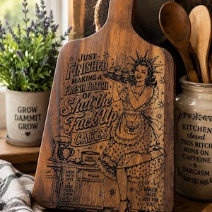 Könnte beinhalten: Ein Schneidebrett aus dunklem Holz mit Griff und einer Schlaufe aus Jutekordel. Das Brett zeigt eine lasergravierte Illustration einer Person in einer Schürze, die ein Tablett mit Cupcakes hält, mit dem Text: JUST FINISHED MAKING A FRESH BATCH OF SHUT THE FUCK UP CAKES.