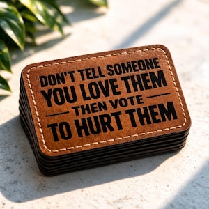 May include: A stack of square brown leather coasters with white stitching around the edges. The coasters have the text "DON'T TELL SOMEONE YOU LOVE THEM THEN VOTE TO HURT THEM" in black.