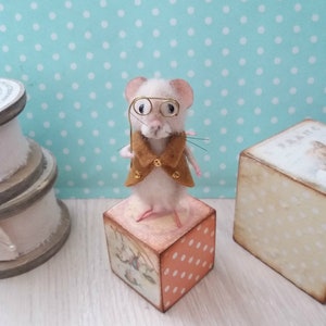 May include: A white felt mouse wearing a brown felt waistcoat and gold-rimmed spectacles, holding a gold wire cane. The mouse is standing on a wooden block with a colourful design.
