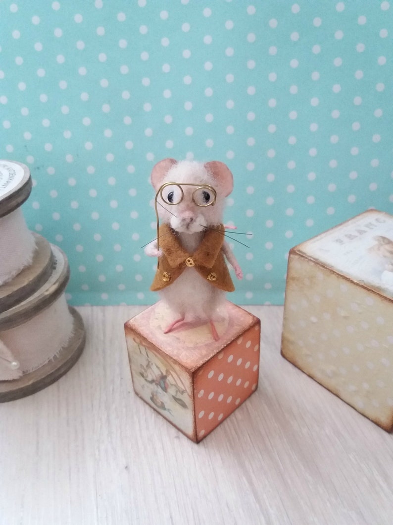 May include: A white felt mouse wearing a brown felt waistcoat and gold-rimmed spectacles, holding a gold wire cane. The mouse is standing on a wooden block with a colourful design.