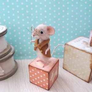 May include: A small, white, needle felted mouse wearing a brown jacket and glasses, standing on a pink and white polka dot wooden block.