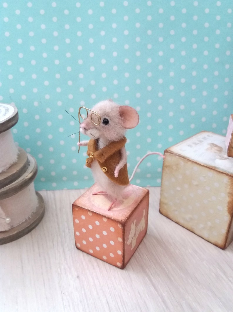May include: A small, white, needle felted mouse wearing a brown jacket and glasses, standing on a pink and white polka dot wooden block.