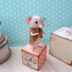 May include: A small, white, needle-felted mouse wearing a brown jacket and gold-rimmed glasses stands on a wooden block with a vintage illustration. The block has a white polka dot pattern on one side.