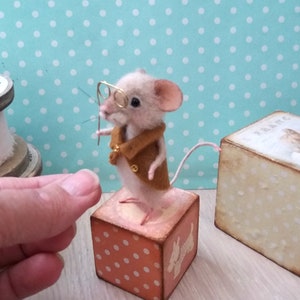May include: A small, beige, needle felted mouse wearing a brown jacket and glasses, standing on a pink and white polka dot wooden block with a white dog silhouette.