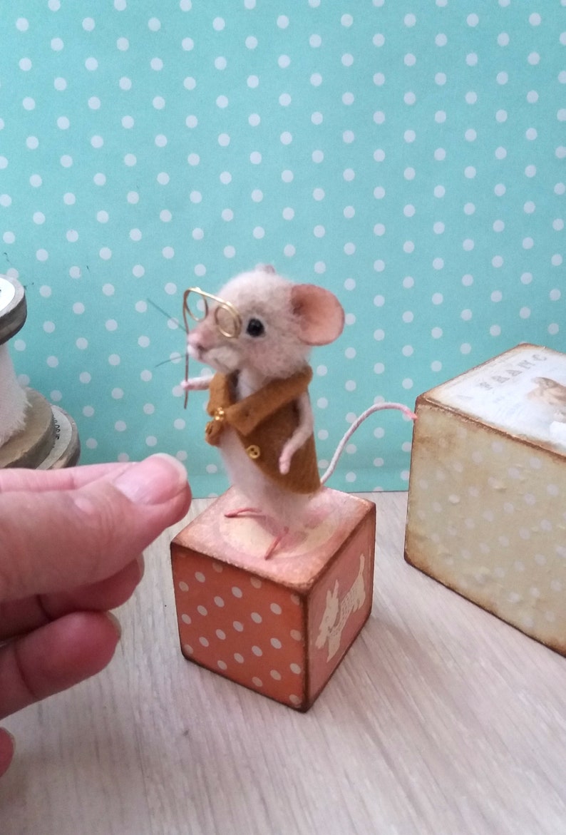 May include: A small, beige, needle felted mouse wearing a brown jacket and glasses, standing on a pink and white polka dot wooden block with a white dog silhouette.