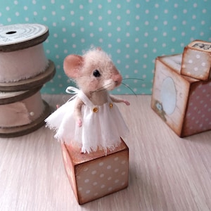Peut inclure: Une figurine de souris en laine feutrée avec une fourrure beige clair, des yeux en perles noires et de longues moustaches. La souris porte une robe blanche sans manches avec de fines bretelles et deux petits boutons dorés sur le corsage.