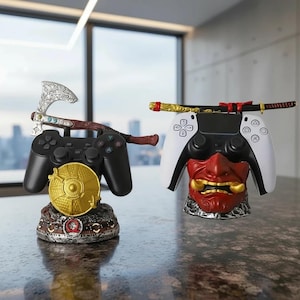 May include: Two video game controller stands. One black controller rests on a stand with a gold disc and axe design. The other white controller rests on a stand with a red demon mask and katana design.