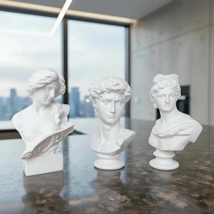 Puede incluir: Tres bustos clásicos blancos de figuras con rasgos faciales detallados y ropa drapeada. Los bustos se exhiben sobre una superficie oscura y texturizada, con un paisaje urbano visible a través de una gran ventana en el fondo. Las esculturas son de diferentes tamaños y estilos.