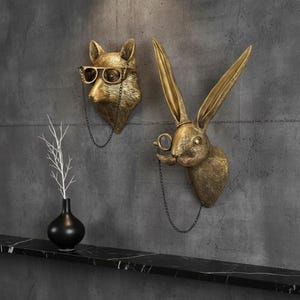 May include: Two gold-coloured animal head wall sculptures: a fox wearing glasses and a rabbit with a monocle. Both have chains attached. A black vase with white branches sits on a black marble shelf.