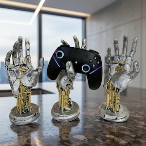 May include: Three robotic hand-shaped game controller stands. The stands are silver and gold with intricate mechanical details. A black game controller with blue accents rests on one stand. The stands are on a brown countertop.