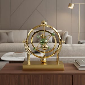 May include: A gold-colored, decorative kinetic sculpture with a spherical design. The sculpture features concentric rings and green glass spheres, mounted on a rectangular base. It is displayed on a wooden surface, with a neutral-toned background.