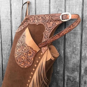 Op de afbeelding: Bruine leren chaps met ingewikkelde bloemmotieven en franjes. De chaps hebben een riem met een zilveren gesp en zijn ontworpen om te paardrijden. Het leer heeft een warme tint en de bloempatronen voegen een decoratief element toe.