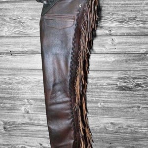 Peut inclure: Chaps en cuir marron avec des franges décoratives sur le côté. Les chaps ont une ceinture noire avec une boucle argentée. Une poche est visible sur le côté. Les chaps sont suspendus sur un fond en bois gris et blanc.
