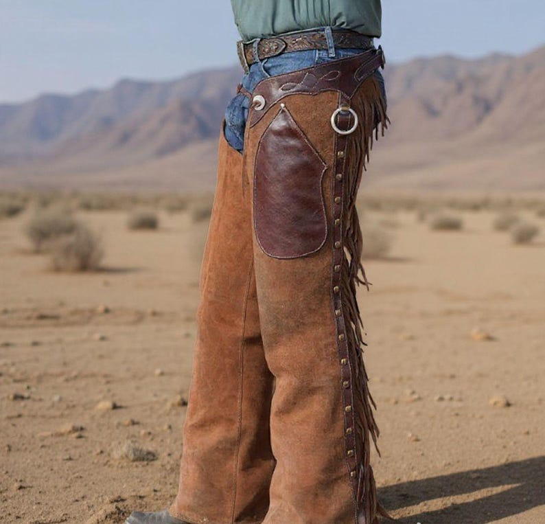 K&ouml;nnte beinhalten: Braune Lederchaps mit einem dunkelbraunen Lederaufn&auml;her am Oberschenkel und Fransen an der Seite. Die Chaps werden mit einem G&uuml;rtel und Jeans getragen, vor einer W&uuml;stenkulisse.