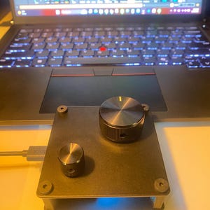 May include: A black square electronic controller featuring one large and one small circular knob on the top surface. The device is secured with four corner screws and is connected to a grey USB cable, with a blue light visible underneath the base.