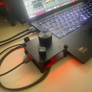 Może przedstawiać: Czarne, prostokątne elektroniczne urządzenie sterujące z dwoma okrągłymi pokrętłami na górnej powierzchni, podłączone kablem USB-C do laptopa. Urządzenie ma matowe czarne wykończenie, widoczne śruby w rogach oraz świecące czerwone światło.
