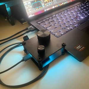 Może przedstawiać: Czarny, niestandardowy panel makro z dwoma okrągłymi pokrętłami na płaskiej, prostokątnej podstawie, podłączony kablem USB-C do laptopa z oprogramowaniem do edycji dźwięku. Urządzenie posiada metalową ramę z widocznymi śrubami w rogach.