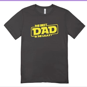 Puede incluir: Una camiseta de manga corta de color gris oscuro con el texto amarillo THE BEST DAD IN THE GALAXY impreso en el pecho. El texto está dispuesto en un formato de bloque estilizado con un contorno amarillo que rodea la palabra DAD y las frases.