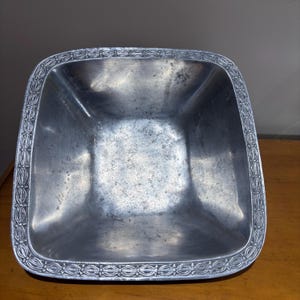 May include: A square metal bowl with a polished silver-toned finish and a textured, repeating decorative border pattern along the rim. The interior surface of the bowl shows a weathered, mottled patina with subtle variations in the metallic sheen.