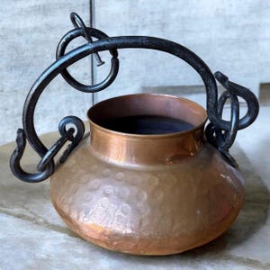 May include: A hammered copper pot with a wide, rounded body and a smooth, cylindrical neck. The pot features a large, arched black iron handle attached to the sides with decorative, curled metal loops that extend outward from the rim.