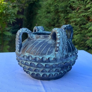 May include: A dark ceramic vessel with a rounded body and four vertical handles. The surface features a textured finish with diagonal grooves on the upper section and repeating scalloped patterns on the lower half, highlighted in teal and black.