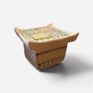 May include: A square ceramic container with a flared, curved rim. The exterior and lid feature a patchwork design of blue and green hand-painted patterns, including swirls, vertical lines, and floral motifs on a cream-coloured glazed surface.