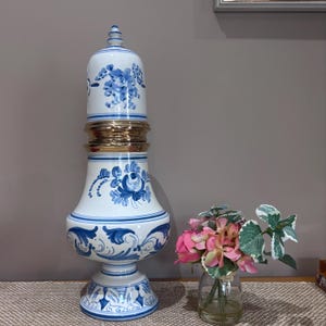 Peut inclure: Un grand vase en céramique blanche orné de motifs floraux bleus et d'une bande dorée métallique au centre. Le vase présente une base arrondie, un milieu effilé et un couvercle bombé. À côté se trouve un petit vase en verre avec des fleurs roses.