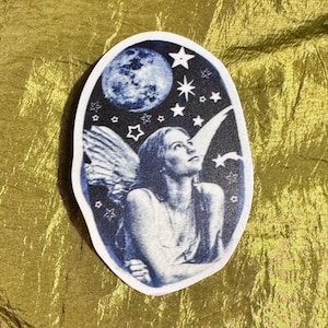 Può includere: Un adesivo di forma ovale con un'illustrazione monocromatica blu e bianca di una persona con grandi ali piumate che guarda verso l'alto. Lo sfondo raffigura un cielo notturno pieno di stelle e una grande luna piena dettagliata.