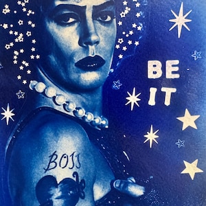 Può includere: Una stampa dai toni blu con il ritratto di una persona dai capelli scuri che indossa una collana di perle. L'immagine include stelle bianche e il testo BE IT. Un tatuaggio sulla spalla mostra la parola BOSS sopra un disegno a forma di cuore.