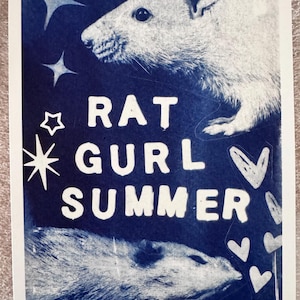 Può includere: Una stampa rettangolare con uno sfondo blu intenso e due illustrazioni di ratti bianchi. Il testo RAT GURL SUMMER è stampato in bianco al centro. Il design include forme di stelle e cuori bianchi che circondano i ratti e il testo.