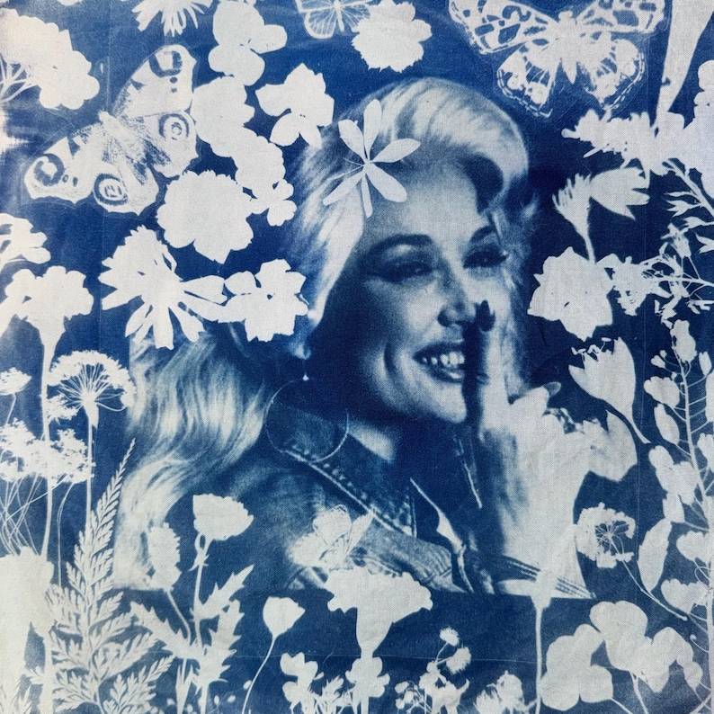 Pu&ograve; includere: Una stampa cianotipica che presenta un ritratto monocromatico blu e bianco di una persona sorridente con i capelli lunghi, circondata da un denso motivo di vari fiori selvatici, felci e farfalle che incorniciano la figura centrale.