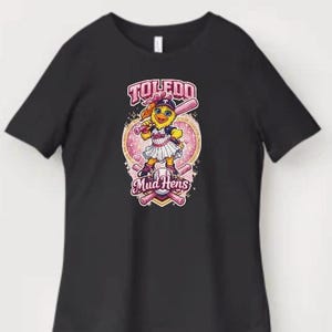 Puede incluir: Camiseta negra con una mascota de dibujos animados de los Mud Hens sosteniendo un bate de béisbol. El diseño incluye el texto "Toledo Mud Hens" en rosa y blanco, sobre un fondo en forma de corazón con bates de béisbol.