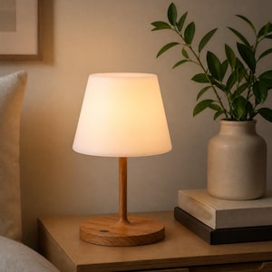May include: A table lamp featuring a smooth, light-colored wooden base and a cylindrical stem that supports a conical, frosted white lampshade. The lamp emits a warm, soft glow and includes a small, circular power button on the wooden base.