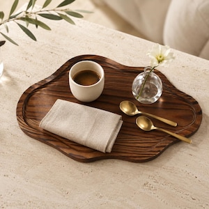 May include: An irregular, dark brown wooden serving tray with a visible wood grain pattern. On the tray sits a matte off-white ceramic cup filled with dark coffee, a folded beige linen napkin, two gold-toned metal spoons, and a small glass vase.