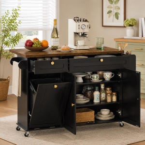 May include: A black kitchen island on wheels with a dark wood top, featuring a pull-out trash bin compartment, two drawers with gold knobs, and open shelving containing various kitchenware, jars, and bottles, plus a side towel rack.