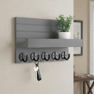 May include: A gray wooden wall-mounted key holder with a shelf and five black metal hooks. A small potted plant sits on the shelf. A key with a black leather fob hangs from one of the hooks. The key holder is mounted on a light-colored wall.