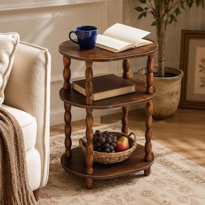 May include: A three-tiered oval wooden side table with turned spindle legs. The top shelf holds a dark blue ceramic mug and an open book. The middle shelf holds a closed book. The bottom shelf holds a woven basket containing grapes and a pear.