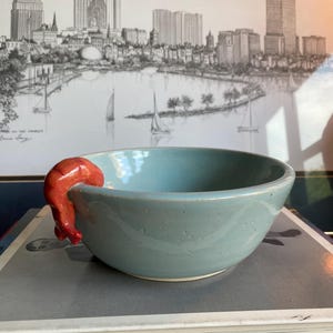 May include: A light blue ceramic bowl with a smooth, speckled finish and a curved rim. A small, bright orange shrimp figurine is attached to the side of the bowl, appearing to hang over the edge. The bowl has a simple, rounded shape.