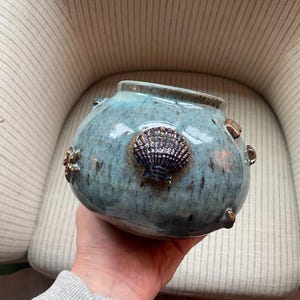 May include: A round ceramic bowl with a glossy blue-green glaze featuring a mottled texture. The exterior is decorated with three-dimensional, dark brown sculpted sea life elements, including a prominent central seashell and smaller starfish.