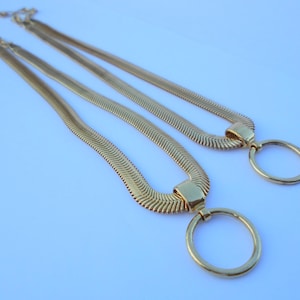 May include: Two gold-colored necklaces with a herringbone chain design. Each necklace features a large, circular pendant. The necklaces are displayed on a light blue surface, creating a contrast with the gold color.