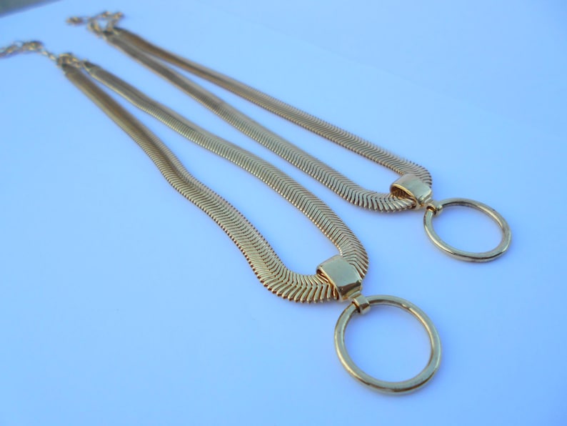 May include: Two gold-colored necklaces with a herringbone chain design. Each necklace features a large, circular pendant. The necklaces are displayed on a light blue surface, creating a contrast with the gold color.