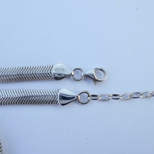May include: A silver-colored necklace with a herringbone chain design. The necklace is made of a shiny metal and features a lobster clasp closure. The clasp is connected to a small chain. The necklace is marked with "925".