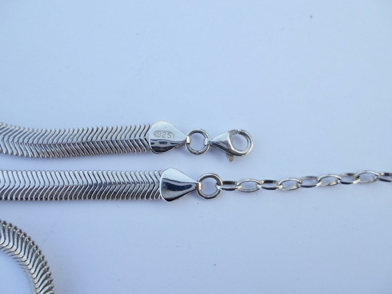 May include: A silver-colored necklace with a herringbone chain design. The necklace is made of a shiny metal and features a lobster clasp closure. The clasp is connected to a small chain. The necklace is marked with "925".