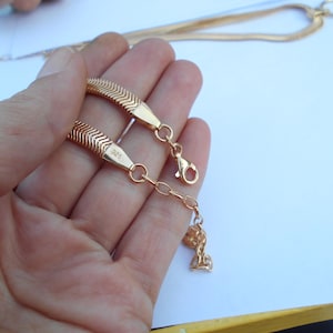 May include: Gold-colored herringbone chain bracelet with a lobster clasp and a small charm. The bracelet is held in a hand against a white background. The clasp is attached to a small chain.