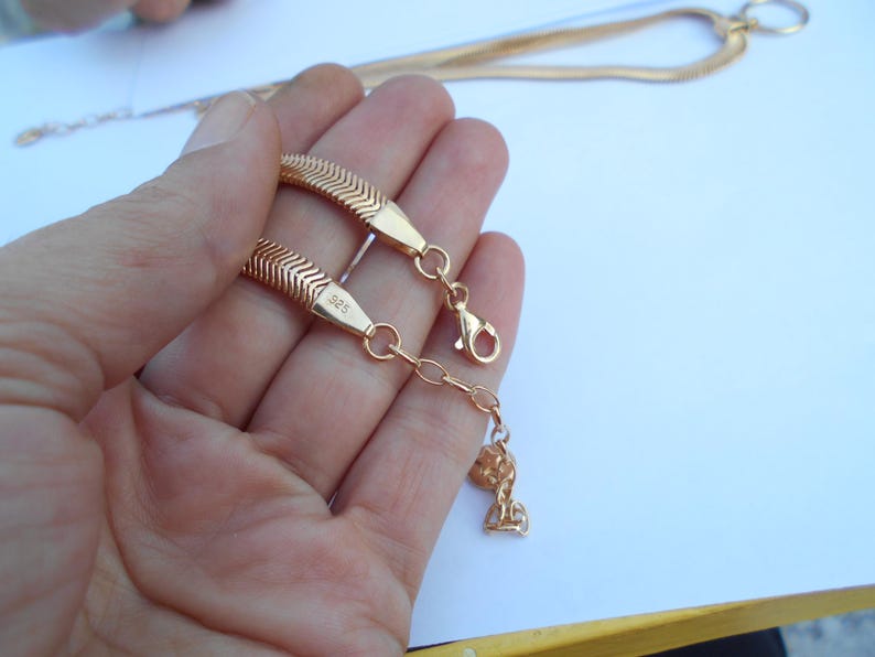 May include: Gold-colored herringbone chain bracelet with a lobster clasp and a small charm. The bracelet is held in a hand against a white background. The clasp is attached to a small chain.
