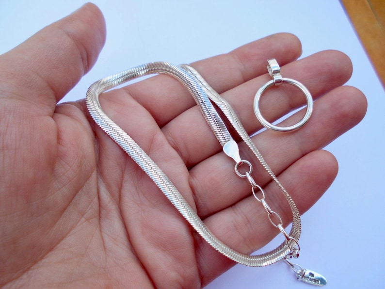 May include: A silver-toned herringbone chain necklace featuring a flat, flexible link design, a circular pendant, and an adjustable extender chain with a small oval tag at the end. The necklace has a polished metallic finish and a lobster claw clasp.