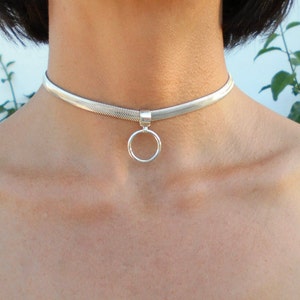 Peut inclure: Un collier ras du cou argenté avec un motif plat et tissé. Un petit pendentif rond est suspendu au centre. Le collier est porté autour du cou.
