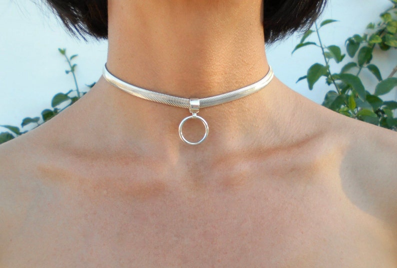 May include: A silver choker necklace with a flat, woven design. A small, round pendant hangs from the center. The necklace is worn around the neck.
